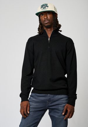 Sweat à zip noir avec manches longues, poignets et ourlet côtelés, porté sur un jean bleu clair, associé à une casquette crème avec des détails graphiques.