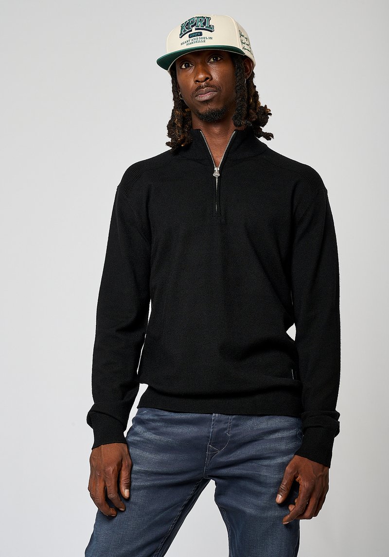 Sweat à zip noir avec manches longues, poignets et ourlet côtelés, porté sur un jean bleu clair, associé à une casquette crème avec des détails graphiques.