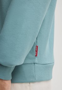 Felpa azzurra con orlo e polsini a coste, dotata di un piccolo tag rosso "JOOP! JEANS" sul lato per il branding. Texture morbida nel complesso.