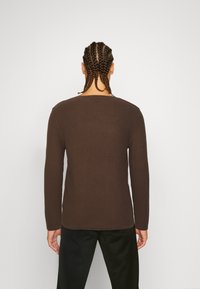 Pull brun à texture gaufrée avec manches longues et large encolure. Le tissu semble doux ; coutures visibles le long des bords et des coutures.