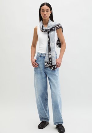 Vrouw die een wit hemdje draagt, lichtblauwe wijde jeans, zwarte loafers en een lange grijze sjaal met bloemenpatroon over haar schouder.