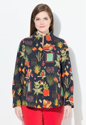 Camisa de botones colorida con patrones florales y de frutas sobre un fondo oscuro, con cuello alzado y mangas largas. Combinada con pantalones rojos.
