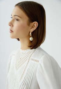 White blouse with vertical pleats, lace detailing, and a scalloped neckline; paired with gold-tone earrings featuring a large round pearl.
