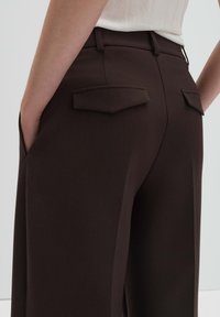 Pantaloni marroni su misura con una texture liscia, dotati di due tasche posteriori con pattine appuntite, tasche laterali e vita aderente.