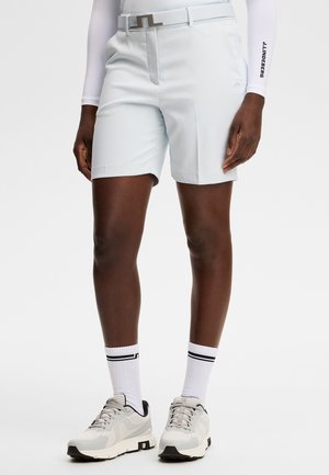 Person iført hvide skræddersyede shorts med bælte, hvid langærmet top, hvide sokker med sorte striber og hvide atletiske sko, der står.