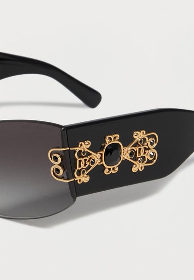 Lunettes de soleil noires avec verres foncés, avec un filigrane doré orné et une pierre noire centrale sur la large branche.