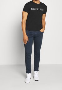 Svart bomullströja med "REPLAY"-logotyp i grått, kombinerad med mörkblå slim-fit jeans och vita sneakers. Enkel, avslappnad klädsel.