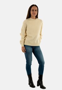 Maglione beige chiaro con maniche testurizzate, polsini e orlo a costine, abbinato a jeans blu scuro e stivaletti neri. Design semplice e aderente.