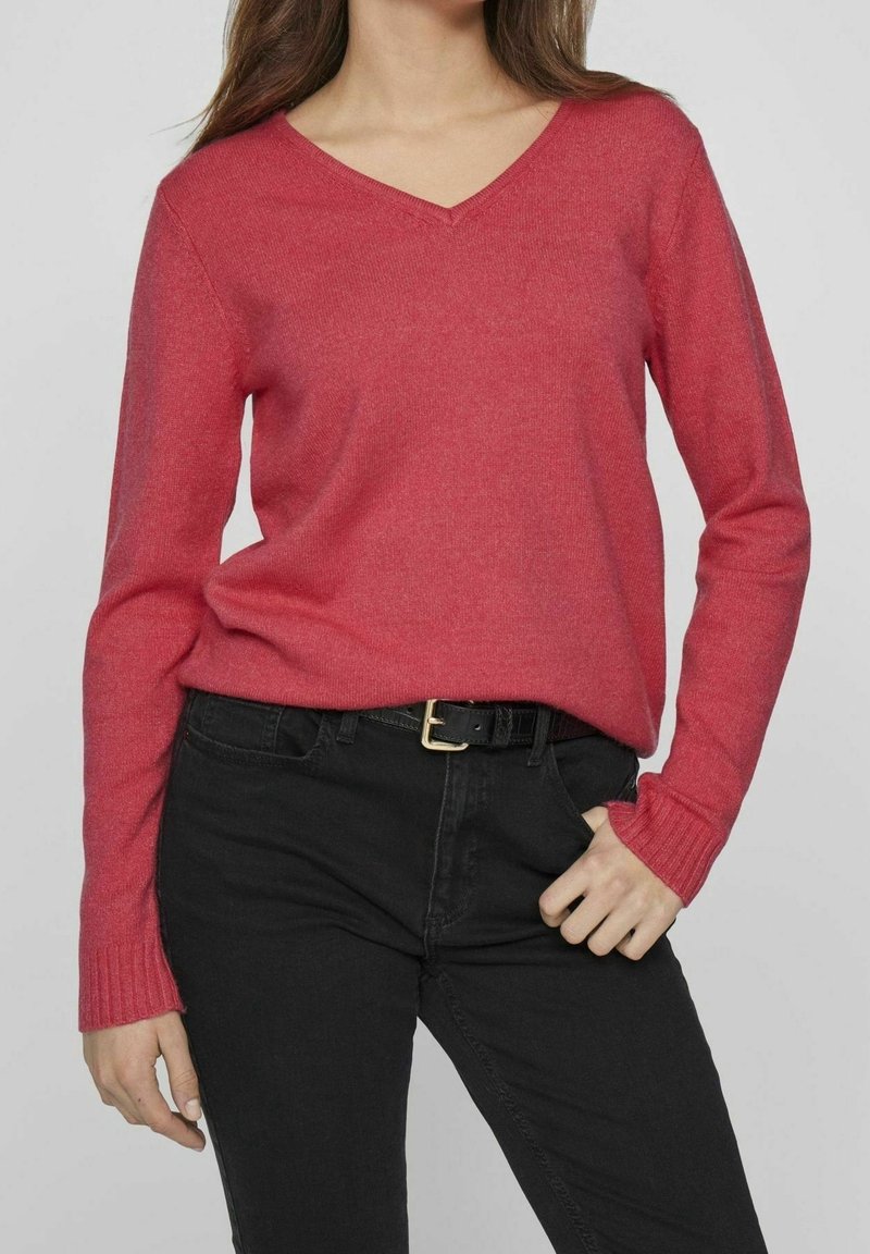 Femme portant un pull rouge à col en V et un pantalon noir avec une ceinture noire, debout avec une main dans la poche devant un fond gris uni.