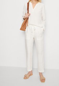 Blouse en lin blanche avec un décolleté en V court, associée à un pantalon en lin blanc à cordon de serrage. Sac à épaule en cuir marron et sandales en cuir tressé beige.