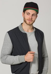 Giacca bomber nera con maniche grigie, dotata di chiusura con zip. Il soggetto indossa una maglietta grigia e un berretto nero con un bordino verde e rosso.
