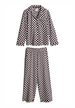 Femme aux cheveux courts, ondulés et foncés, portant une chemise à carreaux noire et blanche boutonnée, regardant pensivement sur le côté sur un fond uni.