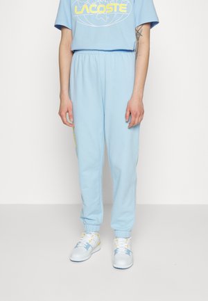 Personne portant un t-shirt Lacoste bleu clair et un pantalon de jogging assorti, associés à des baskets blanches avec des accents bleus et jaunes.