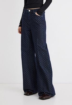 Person iført mørkeblå, viddebukser med høj talje og hvide polkaprikker samt mørke sko med firkantet tå, hånd i lommen.