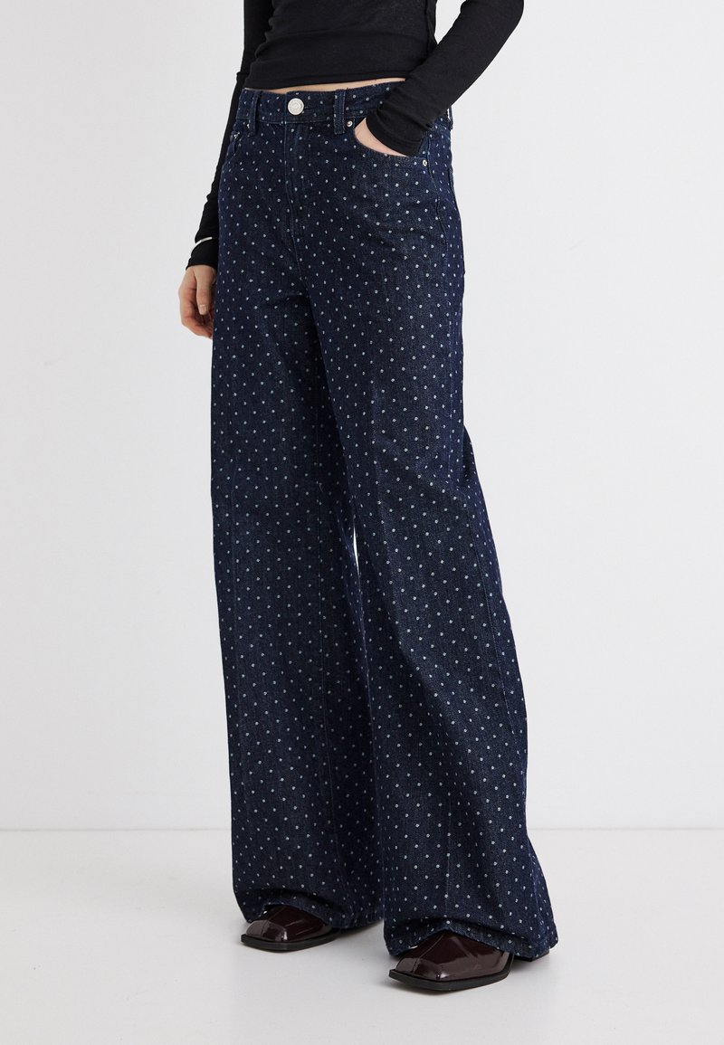 Άτομο που φοράει ψηλόμεσα, φαρδιά σκούρα μπλε τζιν με λευκές πουά και σκούρα παπούτσια με τετράγωνη μύτη, με το χέρι στην τσέπη.