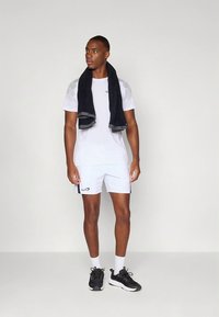 Valkoinen urheiluhihallinen t-paita verkkopaneeleilla, yhdistetty valkoisiin shortseihin, joissa musta logo. Tummanpuhuva pyyhe kaulalla; mustat lenkkarit.