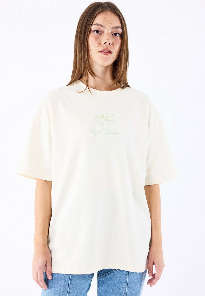 T-shirt surdimensionné de couleur crème en coton, avec des manches courtes et un motif de cœur vert avec un design de carte du monde imprimé sur le devant.