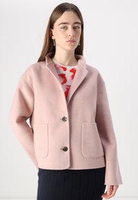 Helle rosa Wollmischung Jacke mit Schalkragen, zwei vorderen Taschen und einem Knopfverschluss. Das Model trägt sie über einem gemusterten Oberteil.