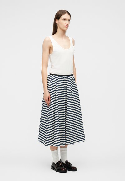 Νεαρή γυναίκα που στέκεται φορώντας λευκό αμάνικο τοπ, μαύρη και λευκή ριγέ φούστα μέχρι το μέσο της γάμπας, λευκές κάλτσες και μαύρα μοκασίνια σε απλό φόντο.