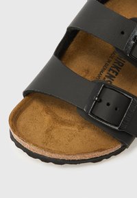 Crne kožne sandale s dva podesiva remena s metalnim kopčama, oblikovanom smeđom brušenom kožom na potplatu i teksturiranim gumenim đonom.