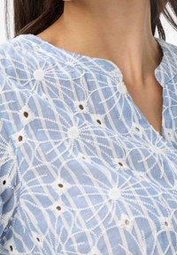 Blouse bleu clair brodée avec des motifs floraux blancs, des rayures horizontales et un col rond avec une patte boutonnée.