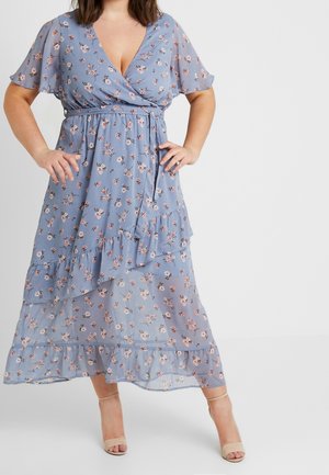 Žena v modrých květinových zavinovacích šatech s krátkými volánkovými rukávy a volánkovým lemem, obutá v béžových otevřených podpatcích, stojící s rukama v bok.