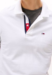 Bílý polo shirt s límcem v námořnické modré, červené a modré pruhy, s knoflíkovou légu a malým vyšitým logem na hrudi.