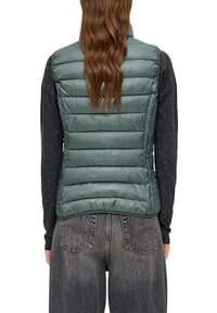 Grön pufferväst med ett quiltat mönster, med hög krage och en slät, glänsande yta. Bärs över en mörk långärmad skjorta.