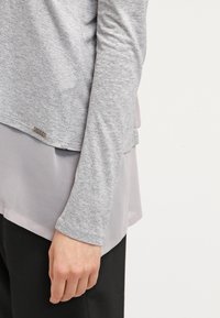 Henkilö, joka käyttää kerroksellista harmaata pitkähihaista puseroa, jossa on läpikuultava helma, ja mustia housuja, käsi rentona sivulla tasaisella taustalla.