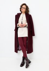 Manteau en laine bordeaux sur un blouse crème avec un devant décoratif, jupe beige, collants bordeaux, et bottines noires brillantes à talon carré.