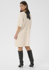 Beige krāsu rotātas kleitas ar apaļu kakla izgriezumu, īsām piedurknēm un melnu lentes dekoru aizmugurē; apvienota ar melnām augstām zābakiem līdz ceļiem.