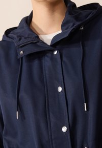 Granatowa kurtka z kapturem, z zapięciem na zamek i zatrzask, kontrastowe białe przeszycia oraz akcenty ze srebrnego metalu. Gładka tekstura.