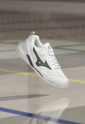 Fehér sportcipő fekete logóval és világoskék fűzőkkel, amely a beltéri sportpálya padlója felett lebeg.