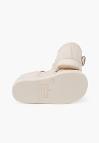 Beige canvas schoenen met een ronde vorm, vastgemaakt met een band en een metalen gesp. Getextureerde rubberen zool met reliëf merkdetails.