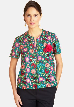 Frau trägt eine grüne Bluse mit kurzen Ärmeln und bunter Blumenstickerei sowie eine rote Blume als Anstecker, kombiniert mit schwarzer Hose und goldenem Schmuck.