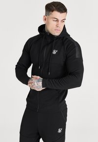 Musta vetoketjullinen huppari, jossa on huppu, tekstitetty muotoilu ja logo rinnassa. Kangas on sileä ja joustava, mikä tekee siitä ihanteellisen aktiiviseen pukeutumiseen.