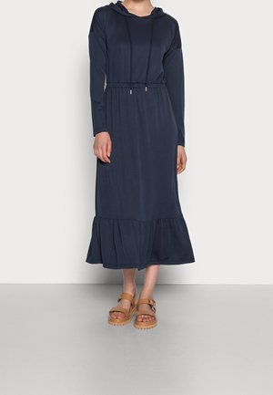 Frau trägt ein langärmliges, marineblaues Midi-Kleid mit Kapuze, geraffter Taille und Saum, kombiniert mit hellbraunen Plateausandalen mit Schnallen.