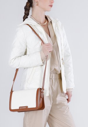 Mujer con chaqueta acolchada blanca y conjunto beige, llevando un bolso de hombro de cuero blanco y marrón con detalles dorados.