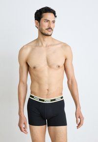 Fekete pamut boxer nadrág széles fehér és fluoreszkáló sárga derékszíjjal, amelyen a "PUMA" márkajelzés látható. A fazon szoros és modern.