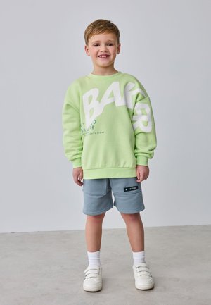 Garçon souriant debout, portant un sweat-shirt vert clair avec un grand texte blanc, un short gris, des chaussettes blanches et des chaussures blanches sur un sol gris.