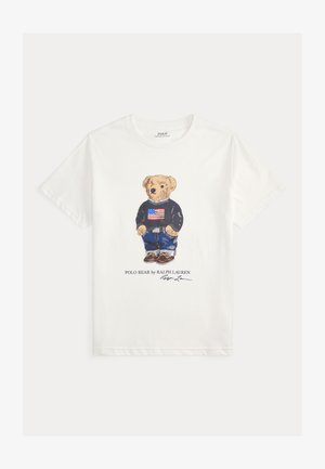 Bela bomba majica iz 100% bombaža s grafikom medveda v modrem puloverju z ameriško zastavo, modrimi kavbojkami in rjavimi čevlji. Pod napisom je tekst "POLO BEAR by RALPH LAUREN."