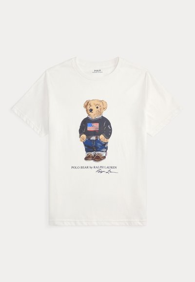 White cotton t-shirt featuring a graphic of a bear in a blue sweater with an American flag, blue jeans, and brown shoes. Text below reads "POLO BEAR by RALPH LAUREN."
