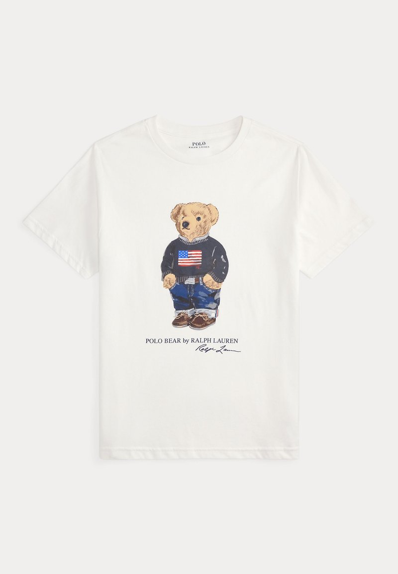Hvit bomull t-skjorte med et grafisk motiv av en bjørn i en blå genser med et amerikansk flagg, blå jeans og brune sko. Teksten under lyder "POLO BEAR av RALPH LAUREN."