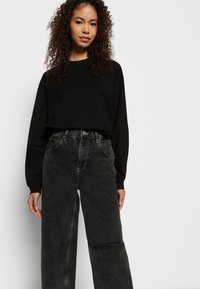 Camisa preta de manga longa combinada com jeans cinzas de cintura alta e desgastados. O tecido parece suave, com um corte relaxado e detalhes de costura visíveis.