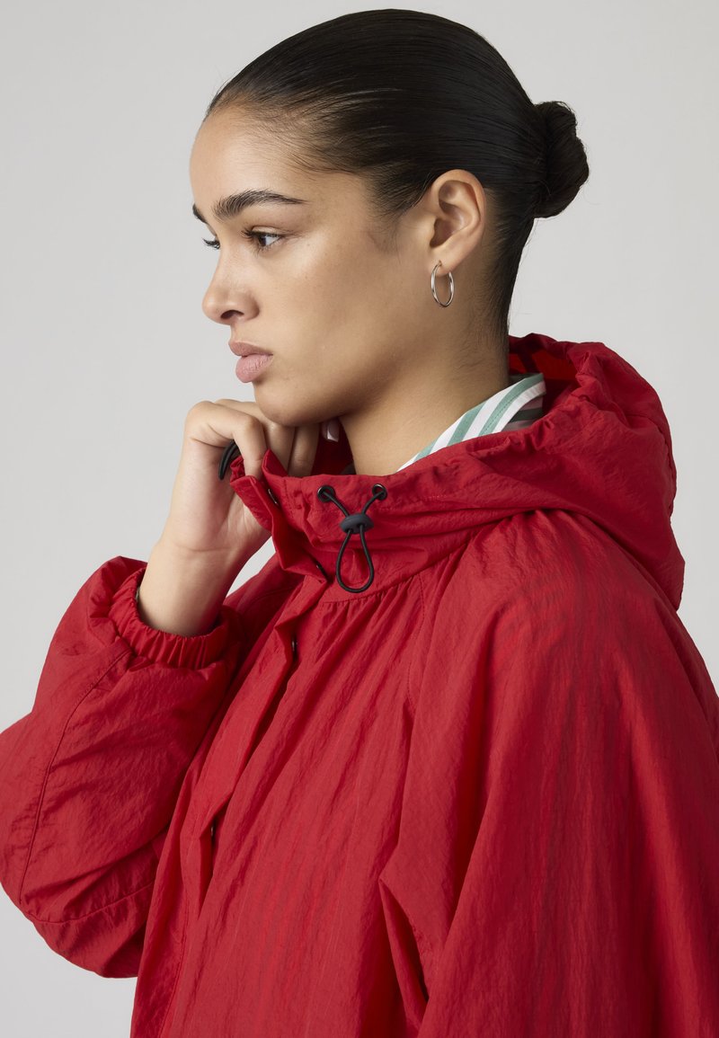 Junge Frau mit dunklen Haaren zu einem Dutt gestylt, die den Kragen einer roten Kapuzenjacke richtet und nachdenklich nach links schaut.