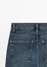 Vista posteriore di jeans in denim blu scuro con due tasche posteriori con dettagli a rivetto e passanti per cintura su sfondo bianco.