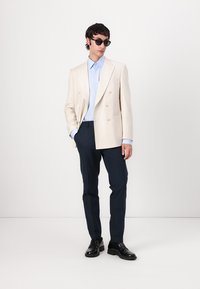 Light beige double-breasted linen blazer over a light blue shirt, paired with dark navy trousers and black leather shoes. Sunglasses included.