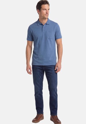 Man die een blauw poloshirt met korte mouwen en een zakje draagt, donkere blauwe jeans en bruine leren sneakers, staand en naar rechts kijkend.