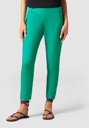 Pantalon slim-fit vert vif longueur cheville, porté avec des sandales noires à lanières et un top noir, montré de la taille aux pieds.