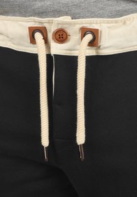 Black pants featuring a cream waistband, adjustable drawstring, and brown leather accents at the eyelets. Smooth fabric texture.